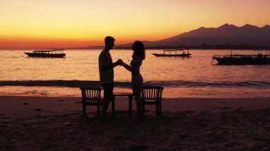Okyanus ve tropik adanın sahillerinde gün batımına karşı duran genç kadın ve erkeğin siluetleri. Birbirine aşık çiftlerin birlikte vakit geçirdiği romantik bir sahne.. 