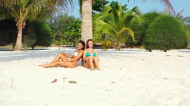 Bikinili iki genç kız arkadaş kumlu deniz kıyısında palmiye ağacının altında güneşleniyor ve konuşuyor. Tropik tatil köyünde dinlenen güzel kadınlar     