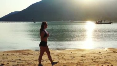 Mayo giymiş çekici genç bir kadın. Güzel adanın okyanusu yakınında dinleniyor, yürüyor ve kulaklıkla müzik dinliyor. 