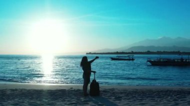 Kadının silueti elinde bavulla sahilde duruyor ve gün batımında akıllı telefon kullanıyor.