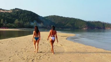 Senkronize bir günde deniz kıyısında yürüyen bikinili iki genç kız arkadaş. Arka manzara Tropik tatil köyünde dinlenen güzel kadınlar    
