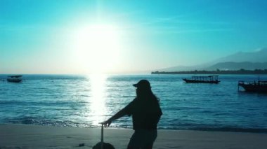Gün batımında elinde bavulla sahilde duran bir kadın silueti.