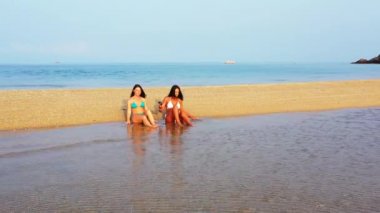 Bikinili iki genç kız deniz kıyısında güneşleniyor ve konuşuyor. Tropik tatil köyünde dinlenen güzel kadınlar    