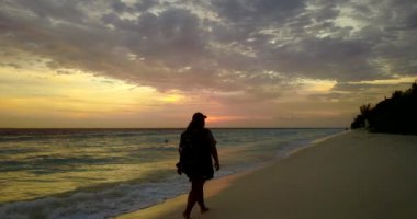 Gün batımında sırt çantasıyla sahilde yürüyen bir kadın silueti.