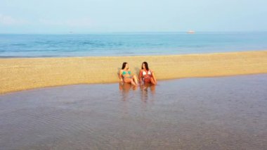 Bikinili iki genç kız deniz kıyısında güneşleniyor ve konuşuyor. Tropik tatil köyünde dinlenen güzel kadınlar    