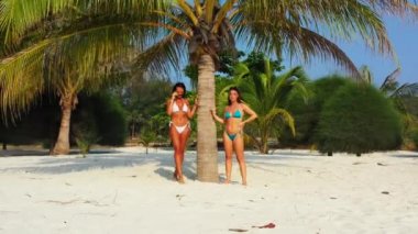 Bikinili iki genç kız arkadaş kumlu deniz kıyısında palmiye ağacının altında güneşleniyor. Tropik tatil köyünde dinlenen güzel kadınlar    