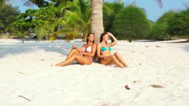 Bikinili iki genç kız arkadaş kumlu deniz kıyısında palmiye ağacının altında güneşleniyor ve konuşuyor. Tropik tatil köyünde dinlenen güzel kadınlar     