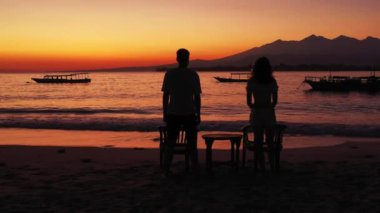 Okyanus ve tropik adanın sahillerinde gün batımına karşı duran genç kadın ve erkeğin siluetleri. Birbirine aşık çiftlerin birlikte vakit geçirdiği romantik bir sahne.. 