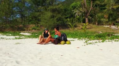 Kumlu deniz kıyısında bavulları yanında oturan ve konuşan iki genç kız arkadaş. Tropik tatil köyünde dinlenen güzel kadınlar   