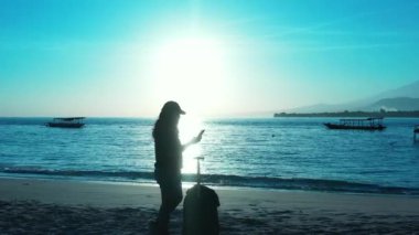 Kadının silueti elinde bavulla sahilde duruyor ve gün batımında akıllı telefon kullanıyor.