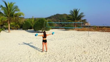 Kadın sporcu kulaklıkla müzik dinliyor ve adanın sahilinde dambıllarla egzersiz yapıyor. 