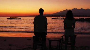 Okyanus ve tropik adanın sahillerinde gün batımına karşı duran genç kadın ve erkeğin siluetleri. Birbirine aşık çiftlerin birlikte vakit geçirdiği romantik bir sahne.. 