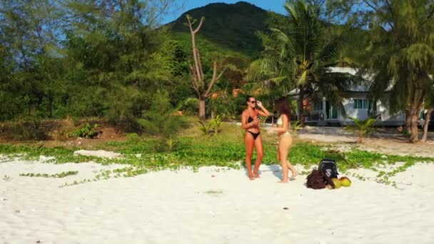 两个穿着比基尼的年轻女友站在沙滩上 一个女孩在手机上为她的女朋友拍照 美丽的女人在热带度假胜地休息 — 图库视频影像