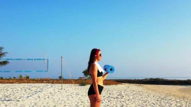 ヘッドフォンで音楽を聞き 島のビーチでダンベルで運動する女性選手 — ストック動画