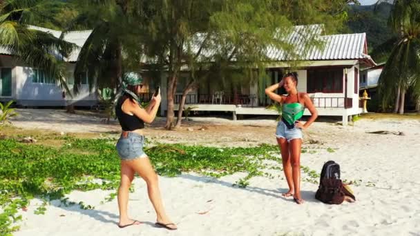 两个年轻的女朋友站在沙滩上 身边背着行李 一个女孩用手机给她的女朋友拍照 美丽的女人在热带度假胜地休息 — 图库视频影像