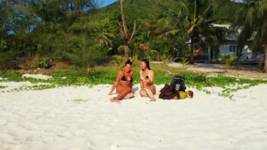 Bikinili iki genç kız arkadaş kumlu deniz kıyısında yatıyorlardı. Bir kız cep telefonu kullanıyor. Tropik tatil köyünde dinlenen güzel kadınlar  