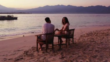 Masanın yanındaki sandalyelerde oturan romantik çift tropikal plajda günbatımının tadını çıkarıyor. 