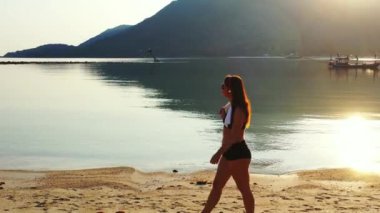Mayo giymiş çekici genç bir kadın. Güzel adanın okyanusu yakınında dinleniyor, yürüyor ve kulaklıkla müzik dinliyor. 