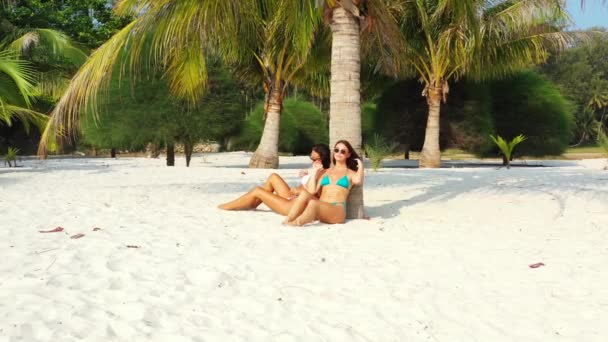 Két fiatal barátnő bikiniben ül a homokos tengerparton a pálmafa alatt, napoznak és beszélgetnek. Gyönyörű nők pihennek trópusi üdülőhelyen     