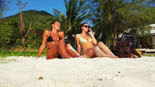 两个穿着比基尼的年轻女友躺在沙滩上 一个女孩在讲手机 美丽的女人在热带度假胜地休息 — 图库视频影像