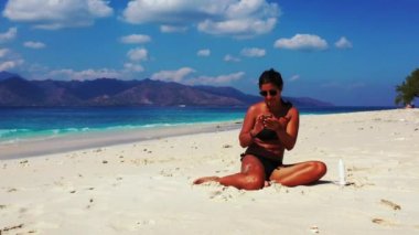 Tropik plajda akıllı telefon kullanan güzel genç bir kadın.