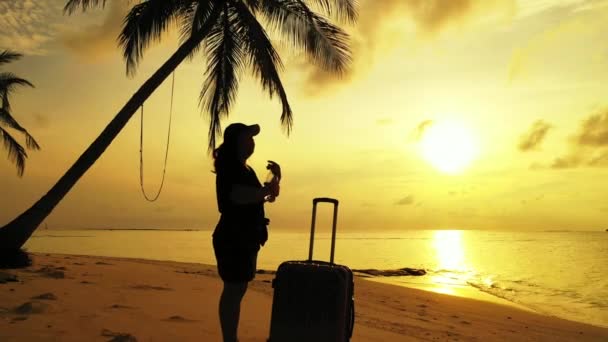 サンセット時にスーツケースを背負ったままトロピカルビーチに立つ女性 — ストック動画