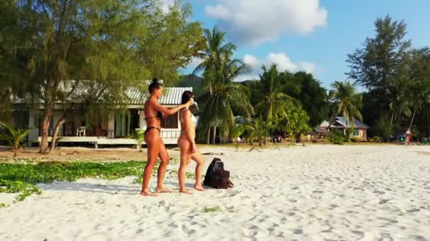 Két fiatal lány barát áll a homokos tengerparton, sundbathing és beszélgetés. Egy lány naptejszínt kent a barátnőjére. Gyönyörű nők pihennek trópusi üdülőhelyen    