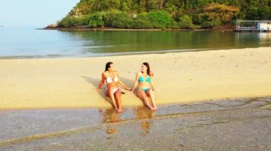 Bikinili iki genç kız deniz kıyısında güneşleniyor ve konuşuyor. Tropik tatil köyünde dinlenen güzel kadınlar    