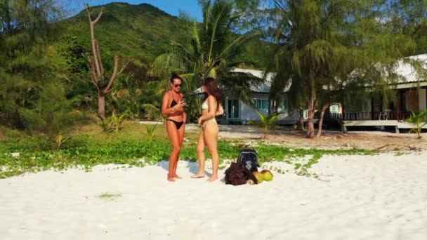 两个穿着比基尼的年轻女友站在沙滩上 用手机自拍 美丽的女人在热带度假胜地休息 — 图库视频影像