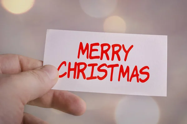 Cartão na mão com a palavra Feliz Natal no fundo do bokeh — Fotografia de Stock