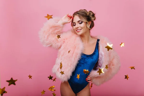 Cheerful curly girl in velvet bodysuit posing with eyes closed standing on pink background. Charming young woman fur coat celebrating her birthday, having fun with golden star confetti and smiling.