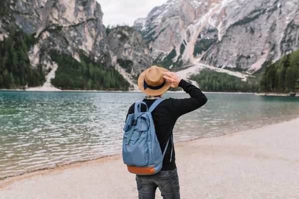 Stylish guy wearing vintage hat decorated with ribbon relaxing on the lake shore and looking at water. Portrait of young man with travel backpack enjoying mountain landscape in cloudy morning..