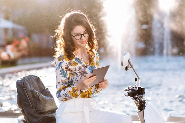 Cute brunette student does her homework outdoor chilling next to city fountain after lessons. Portrait of reading girl with gadget resting in the park near scooter and waiting friends..