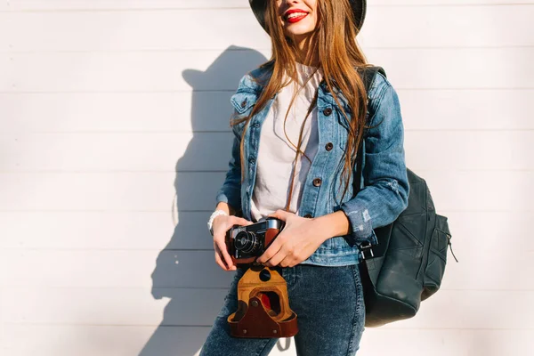 Smiling female photographer in jacket standing in front of wall ready to make new photo. Adorable young brunette woman in trendy jeans outfit and hat posing isolated on wooden background with camera.