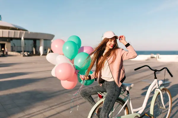 Smiling happy girl with long hair waiting friends at the sea pier for a joint bike ride in morning. Portrait of cheerful brunette young woman with bicycle and party balloons having fun at the ocean.