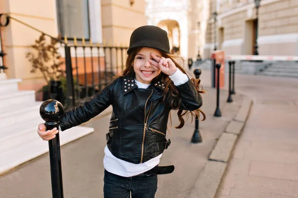 Portrait of laughing little girl in trendy black jacket and white shirt waiting friends on the street. Cute long-haired kid with amazing smile posing outside on city background after lessons.