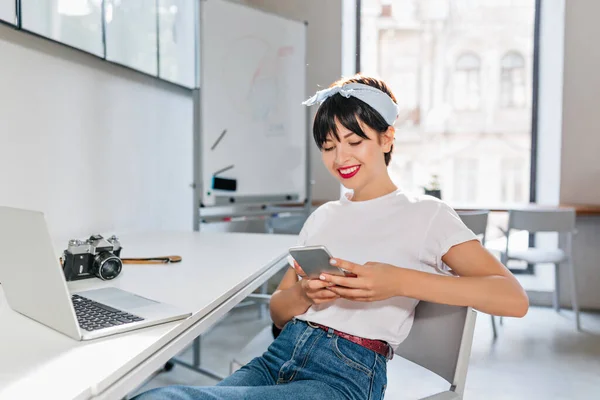 Joyful brunette lady in white shirt and blue jeans working with laptop in big modern office. Pretty girl with short hairstyle texting message while resting at the table with computer and camera on it