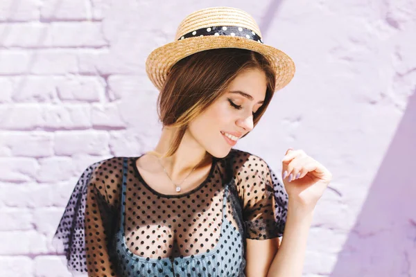 Lovely lady wears vintage hat and silver necklace looking away while posing near old white wall. Outdoor portrait of shy smiling girl with pale skin standing with eyes closed, enjoying sunlight..