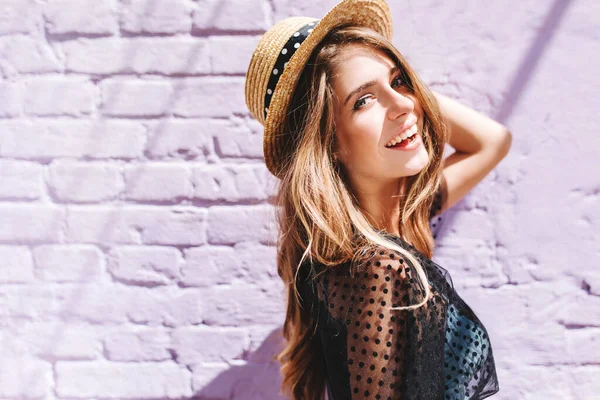 Graceful young woman with long shiny hair gladly posing while walk outside in good mood. Outdoor close-up photo of pleased girl in trendy summer hat with black ribbon enjoying sunshine..