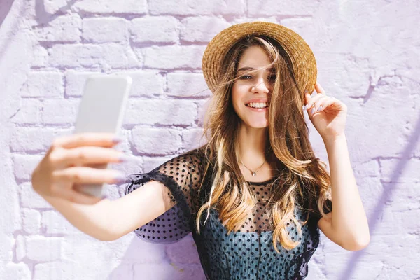 Charming girl with elegant necklace making selfie in front of old white wall. Outdoor close-up portrait of laughing blonde young lady in straw hat holding smartphone and taking picture of herself..