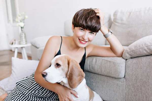 Wonderful tanned girl propping head with hand posing in home after funny game with beagle dog. Indoor close-up portrait of pretty happy woman with short hair sitting on the floor with puppy..