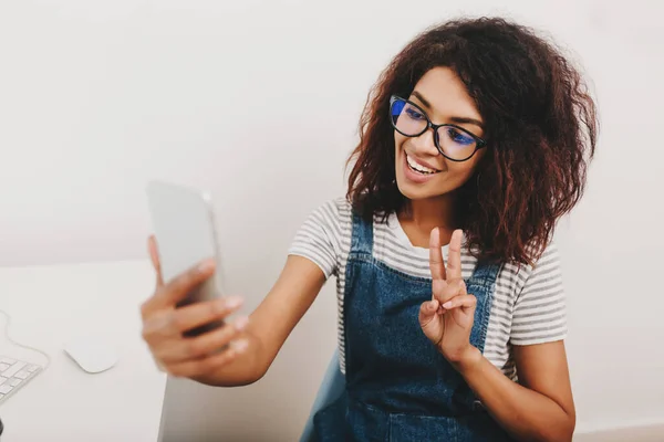 Excited black girl with pretty hairstyle taking picture of herself resting from work. Charming smiling young lady wearing glasses making selfie using white smartphone isolated on gray background..