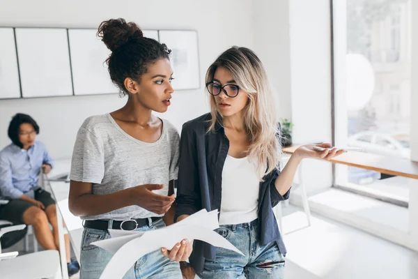 African female office worker and her blonde colleague discussing financial report. Indoor portrait of two young business-ladies talking about job and asian man on background..