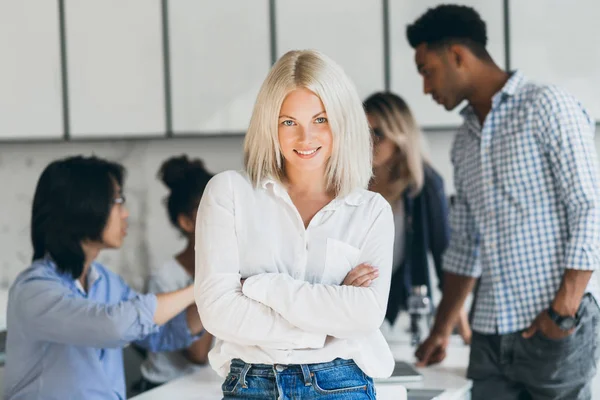 Stylish female secretary standing in confident pose in conference hall and smiling. Indoor portrait of pretty blonde office worker waiting for negotiation with partners..