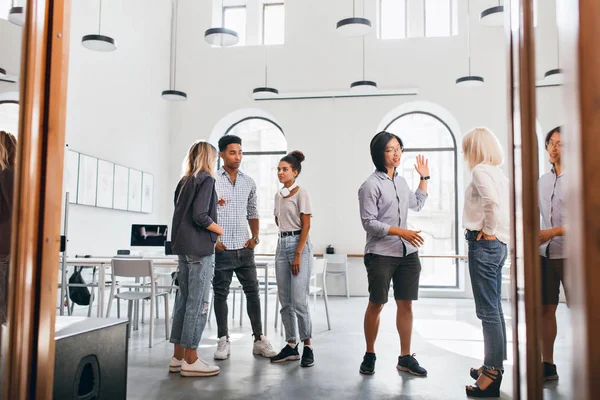 Asian student in shorts and black sneakers talking with blonde female friend while waiting for lecture. Indoor full-length portrait of african boy and girl standing in university hall..