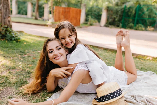 Glad young woman with sparkle make-up chilling on blanket with legs up and smiling to camera. Laughing tanned girl lying on mothers back during photoshoot in park..