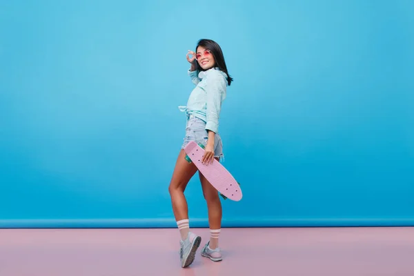 Full-length portrait of ecstatic asian girl looking over shoulder while posing with skateboard. Indoor photo of slim latin girl in denim shorts standing in confident pose on blue background..