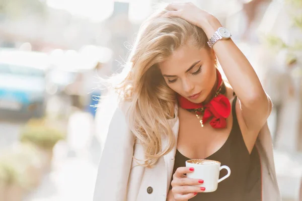 Tired blonde girl touching her hair while holding cup of cappuccino on blur background. Dreamy young woman in coat standing with eyes closed enjoying coffee in sunny morning..