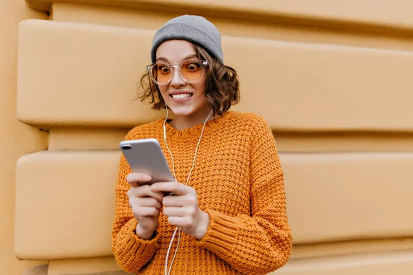 Pleased female model making funny faces while talking with friend on video call. Outdoor portrait of joyful curly girl in yellow sweater and gray hat standing on the street with phone..