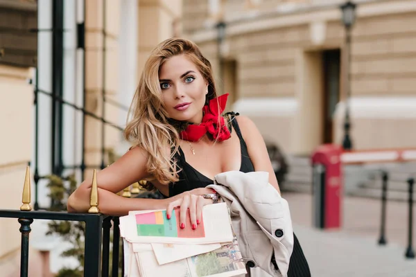 Surprised blue-eyed female model in golden pendant leaning to iron fence on blur background. Shocked fair-haired woman gently smiling while standing on the street with newspapers and coat in hands..